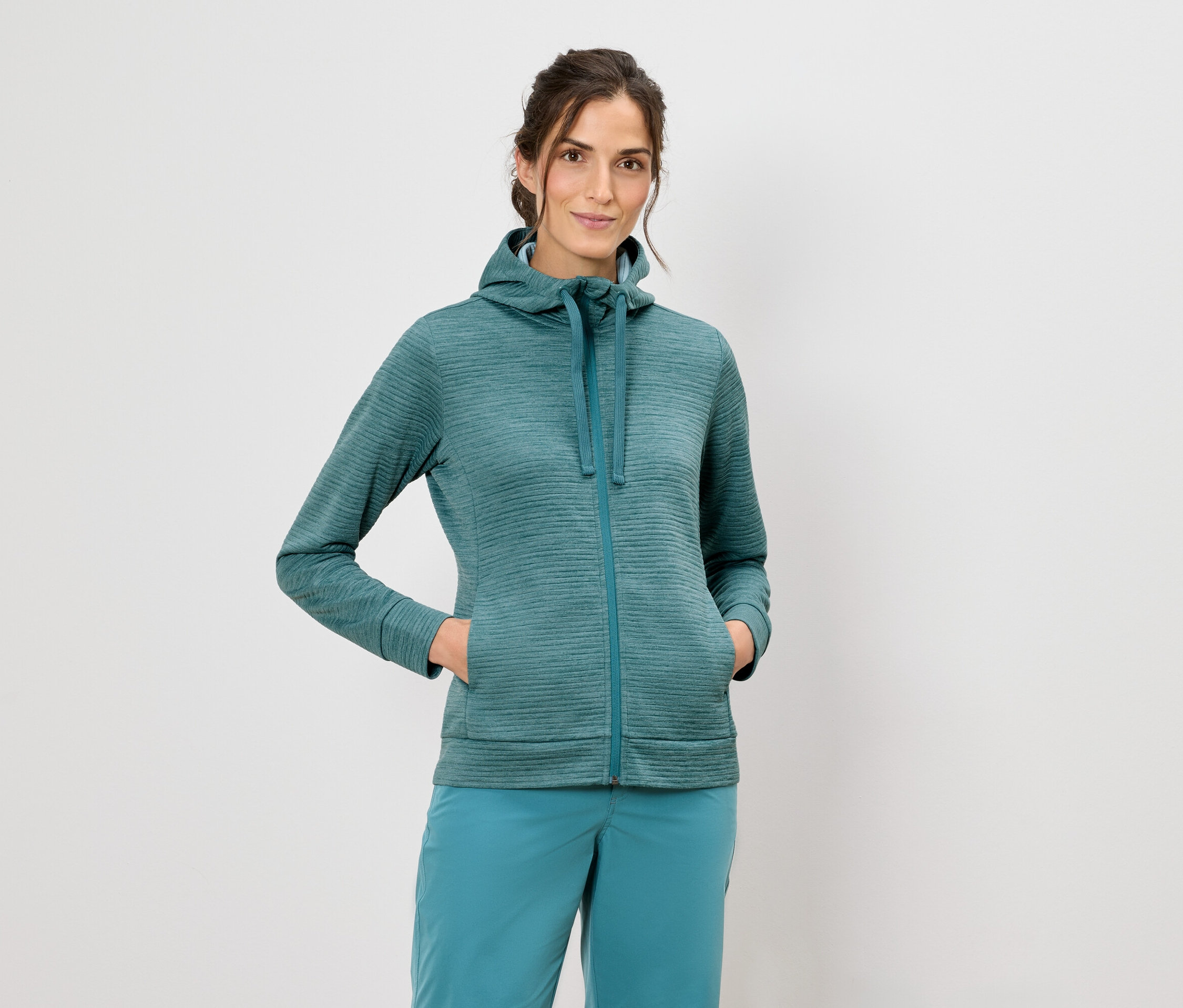 Une femme portant un sweat à capuche et un pantalon bleu se tient devant un fond blanc.