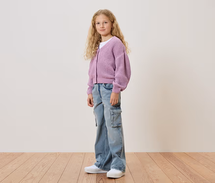 Une fille aux cheveux blonds et bouclés porte un cardigan violet, un jean et des chaussures blanches.