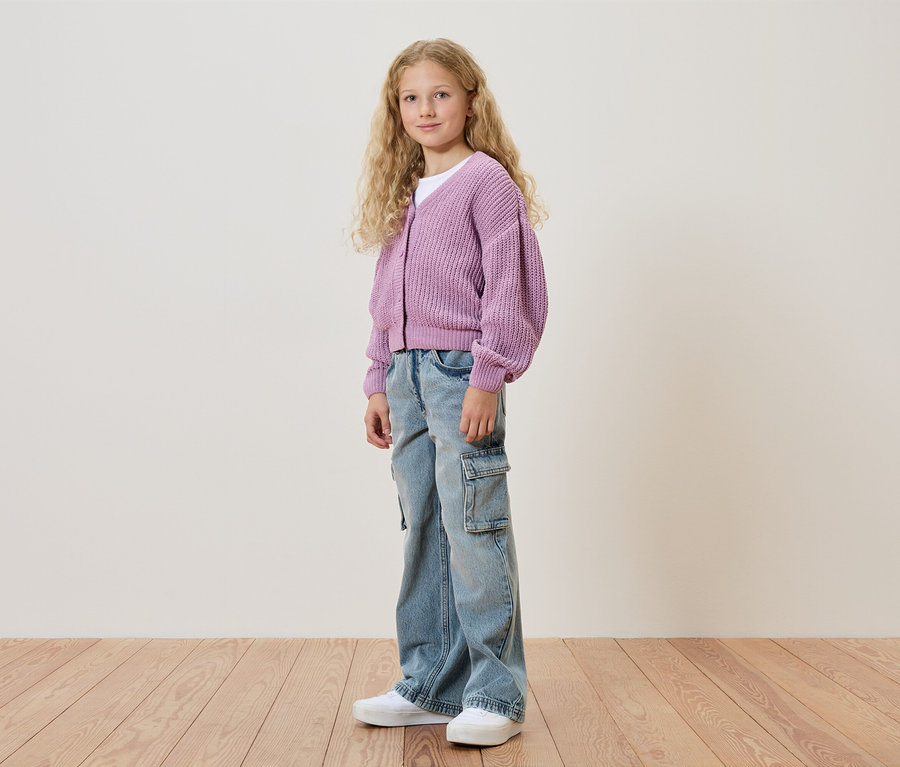 Une fille aux cheveux blonds et bouclés porte un cardigan violet, un jean et des chaussures blanches.