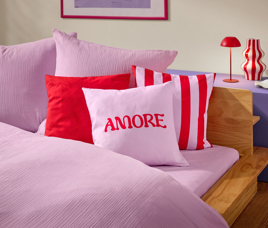 Gros plan d'un lit avec des draps et des oreillers roses. Un oreiller porte le mot AMORE en rouge.