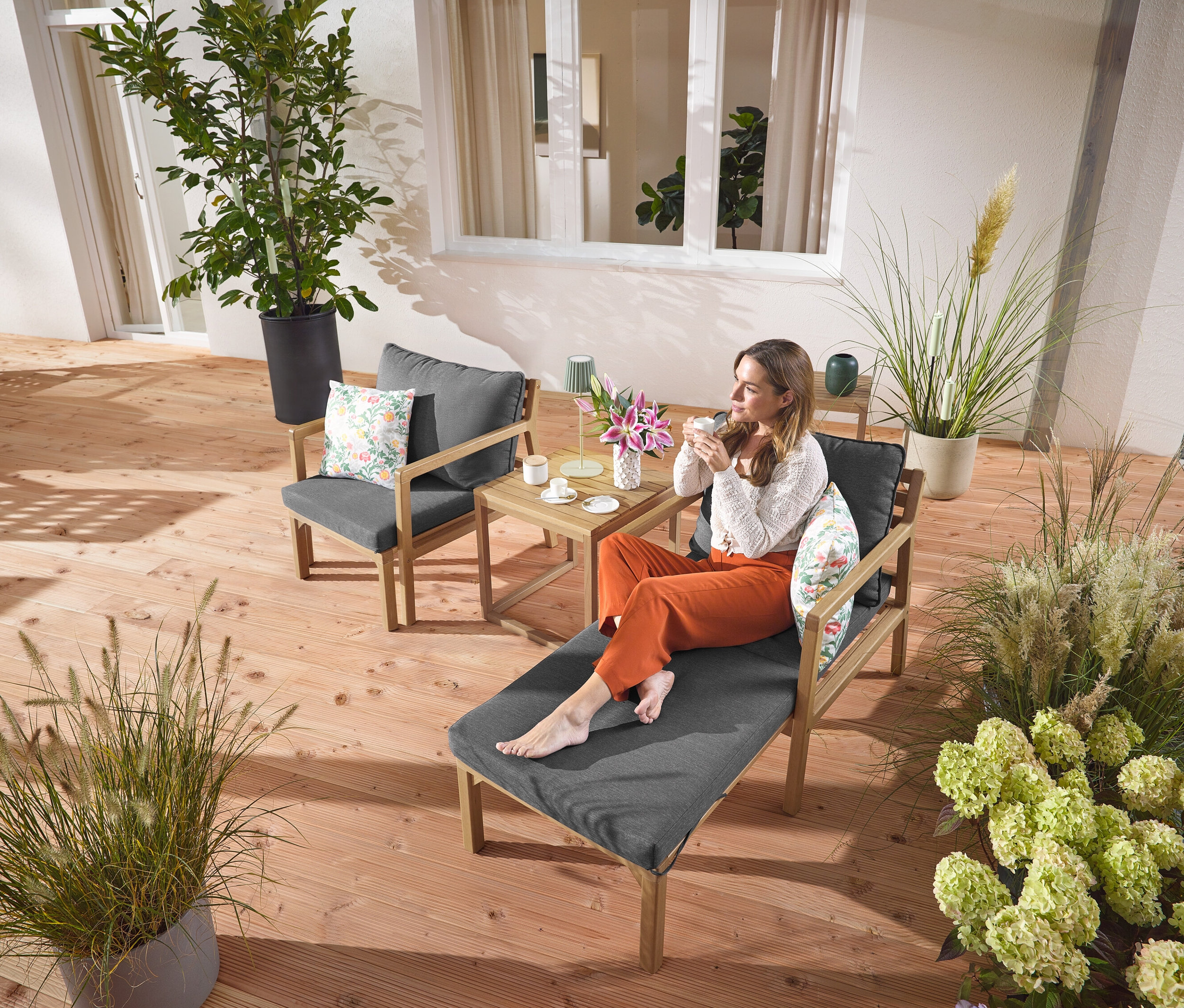 Une femme est assise sur un fauteuil de jardin extensible avec une boisson. Une lampe de table d’extérieur à LED rechargeable sauge est posée sur une table à côté d’elle. À l’arrière-plan, on voit 2 coussins d’extérieur avec motif floral.