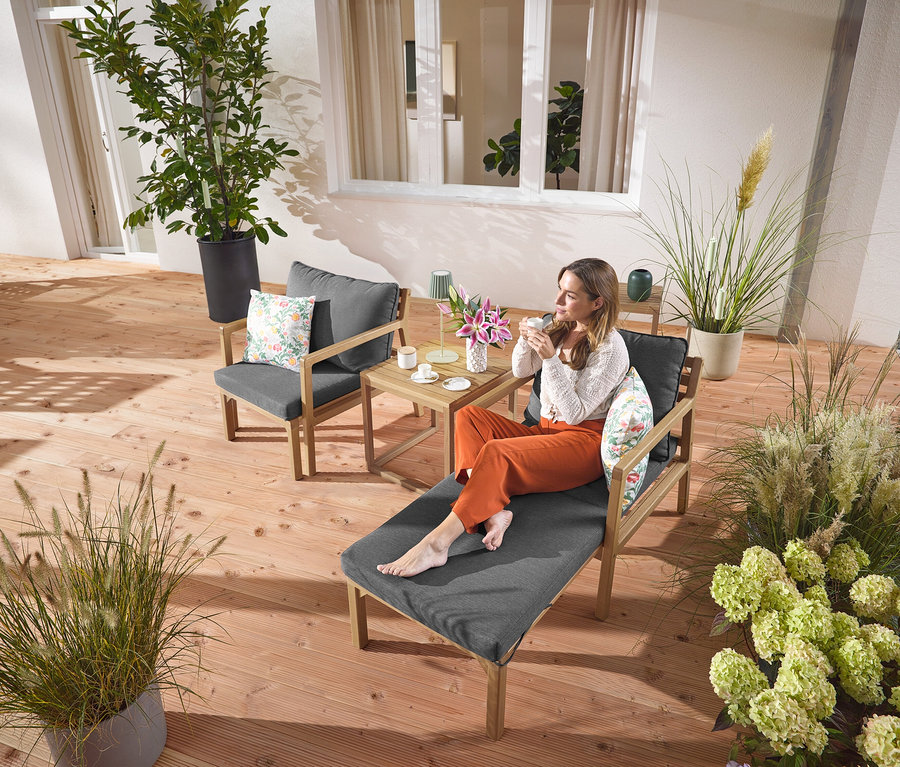 Une femme est assise sur un fauteuil de jardin extensible avec une boisson. Une lampe de table d’extérieur à LED rechargeable sauge est posée sur une table à côté d’elle. À l’arrière-plan, on voit 2 coussins d’extérieur avec motif floral.