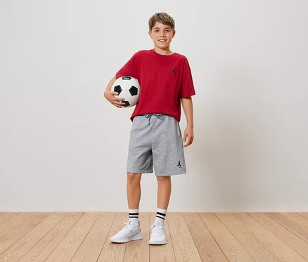 Un garçon souriant tient un ballon de foot dans ses bras, il porte un t-shirt rouge et un short gris.