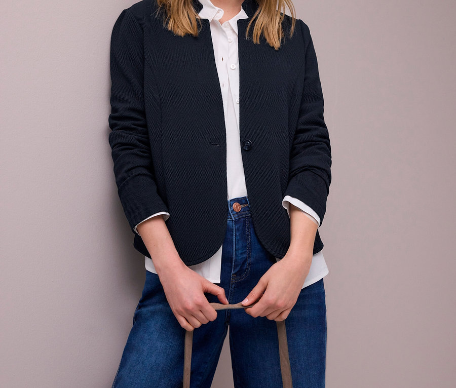 Femme blonde portant un blazer en jersey Street One bleu foncé, une chemise blanche et un jean bleu, tenant un sac à main marron clair.