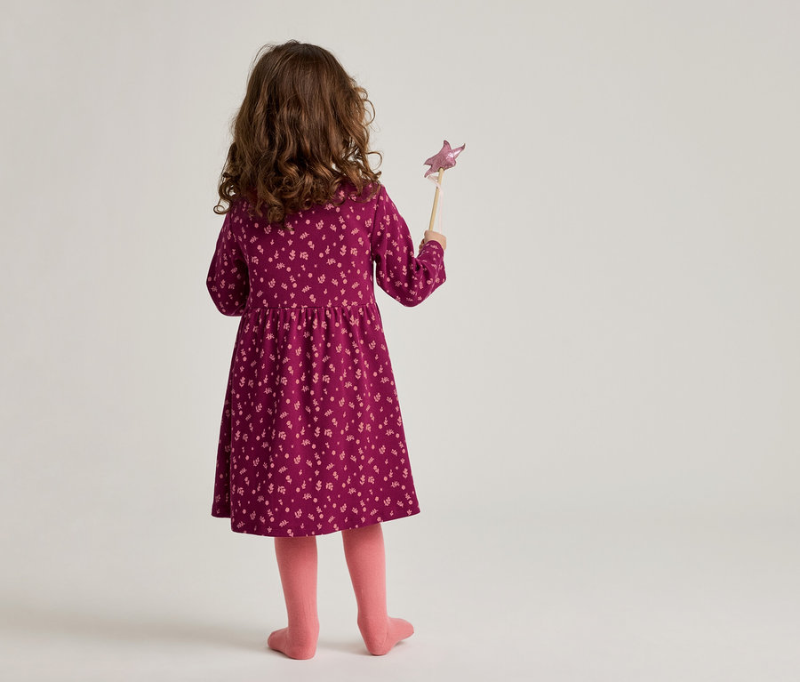 Vue arrière d'une fillette aux cheveux bruns et bouclés portant une robe bordeaux à fleurs et des collants roses, tenant une baguette magique.