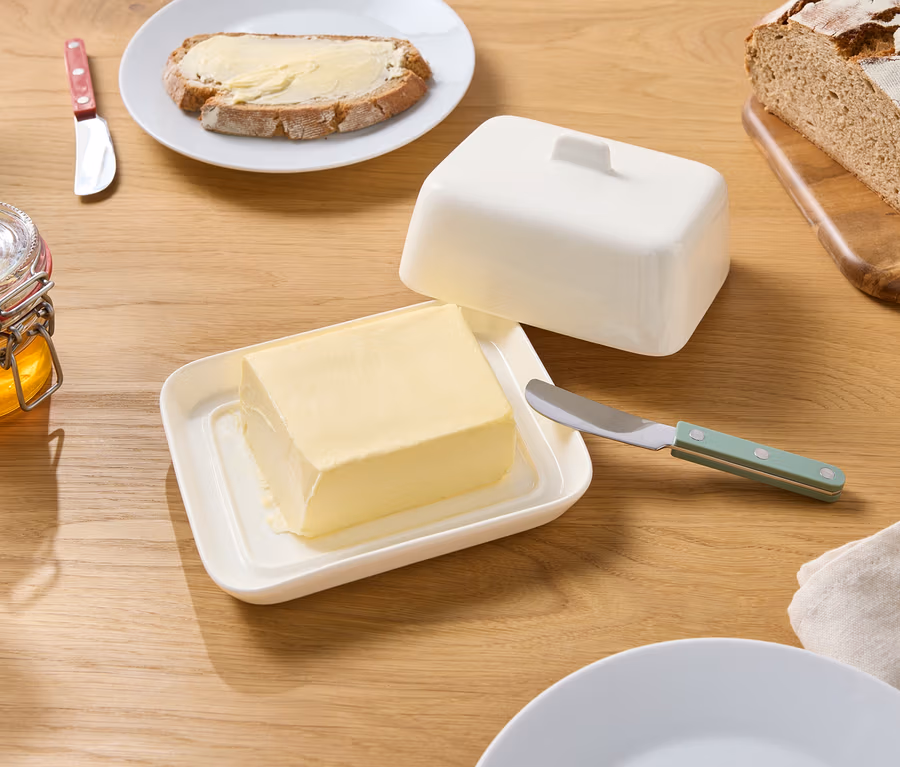Sur une table en bois, il y a un beurrier ouvert avec du beurre, un pot de miel, un couteau, du pain et une assiette avec une tartine beurrée.