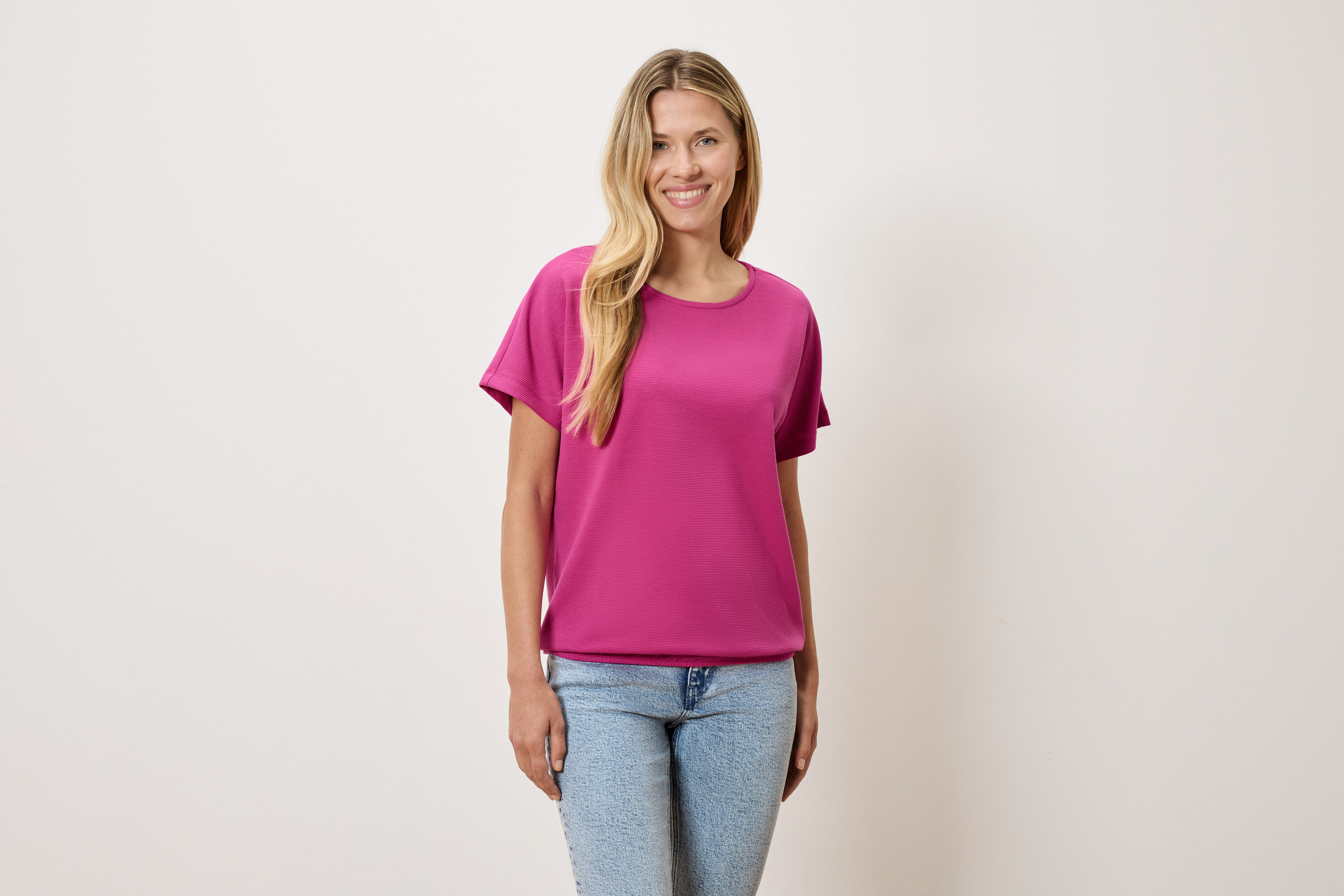 Une femme aux cheveux blonds porte un t-shirt rose et un jean bleu.