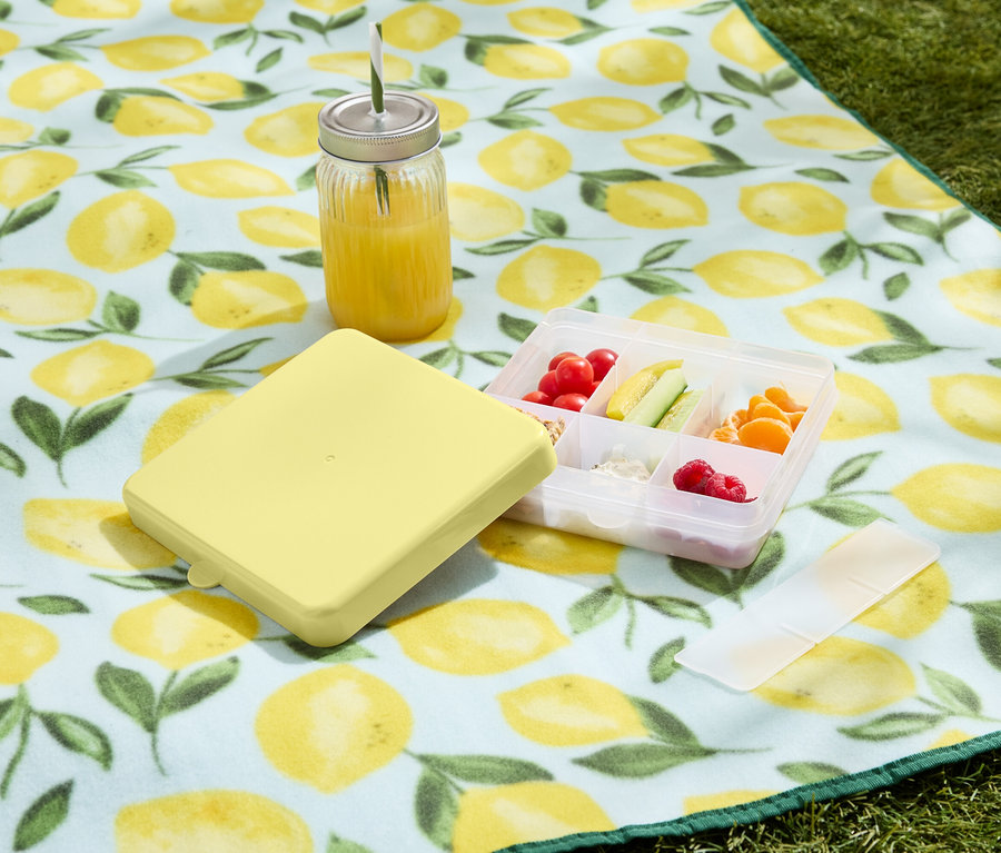 Boîte à lunch jaune avec récipient rempli de fruits et légumes et boisson sur une couverture de pique-nique à motif citron.