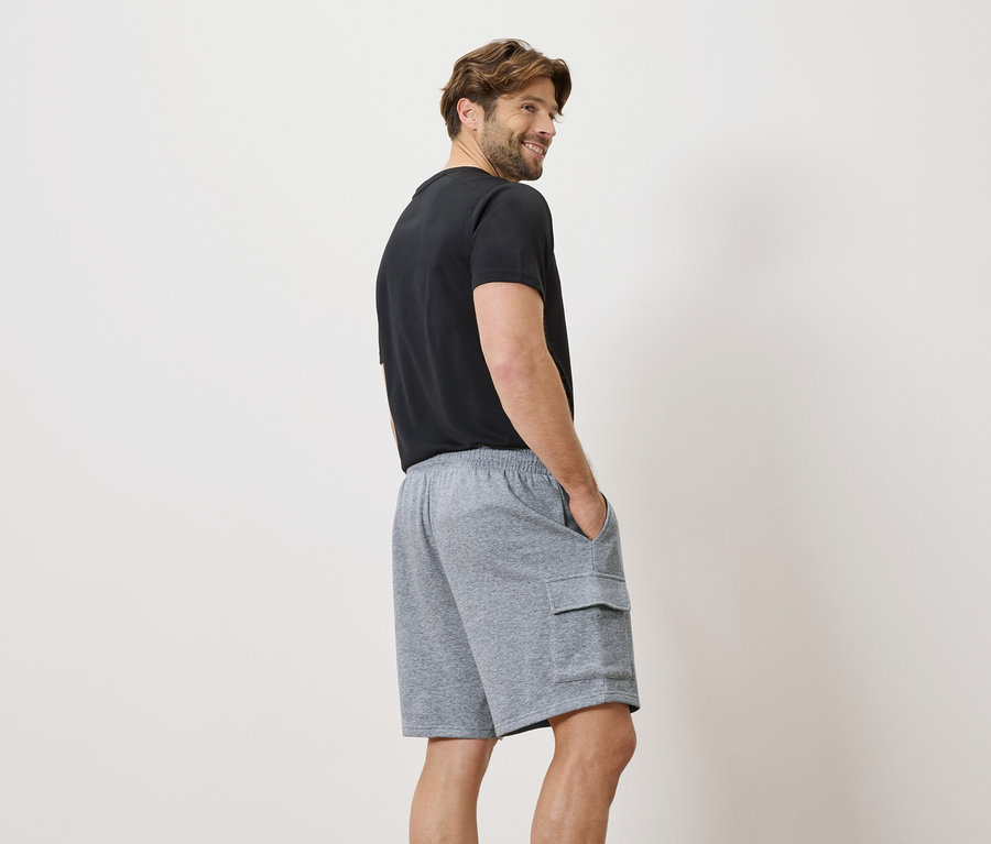 Homme portant un t-shirt noir et un short cargo gris, les mains dans les poches, regardant par-dessus son épaule.