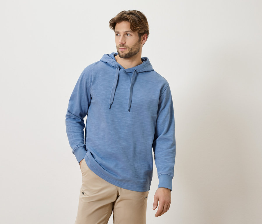 Homme barbu portant un sweat à capuche bleu avec cordon de serrage et un pantalon beige, les mains dans les poches.
