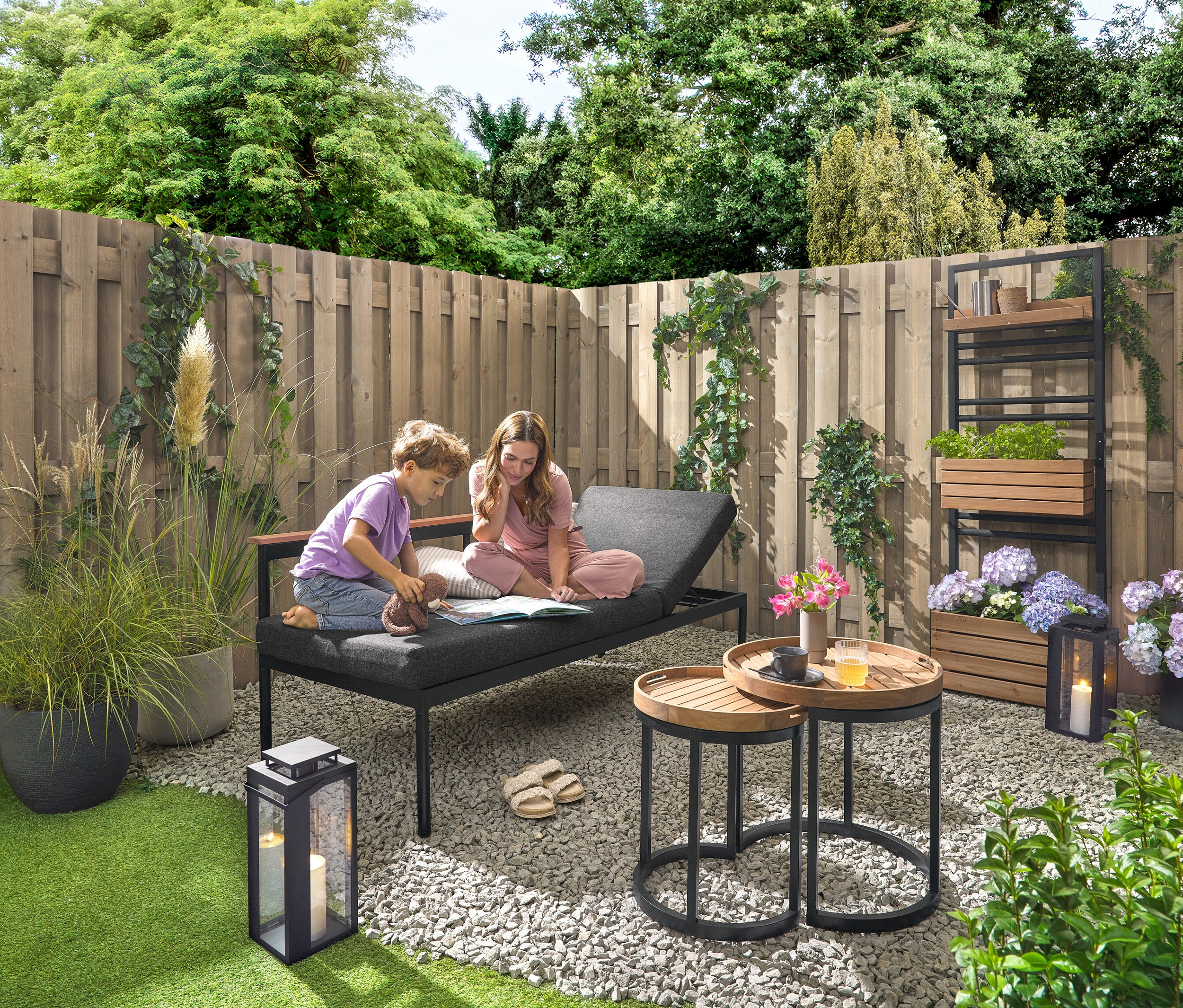Un enfant et une femme sont assis sur un banc de jardin 2 en 1 ‒ Chaise longue «
Aline
A» et lisent un livre. Un mur végétal avec des étagères flexiblement accrochables «
Aline
A» se trouve à l’arrière-plan. Une lanterne d’extérieur avec bougie se trouve à droite. Deux tables d’appoint de jardin avec plateaux amovibles, Ø env. 40
Acm et 50
Acm sont également disponibles, ainsi que 2
coussins d’extérieur avec toucher lin.