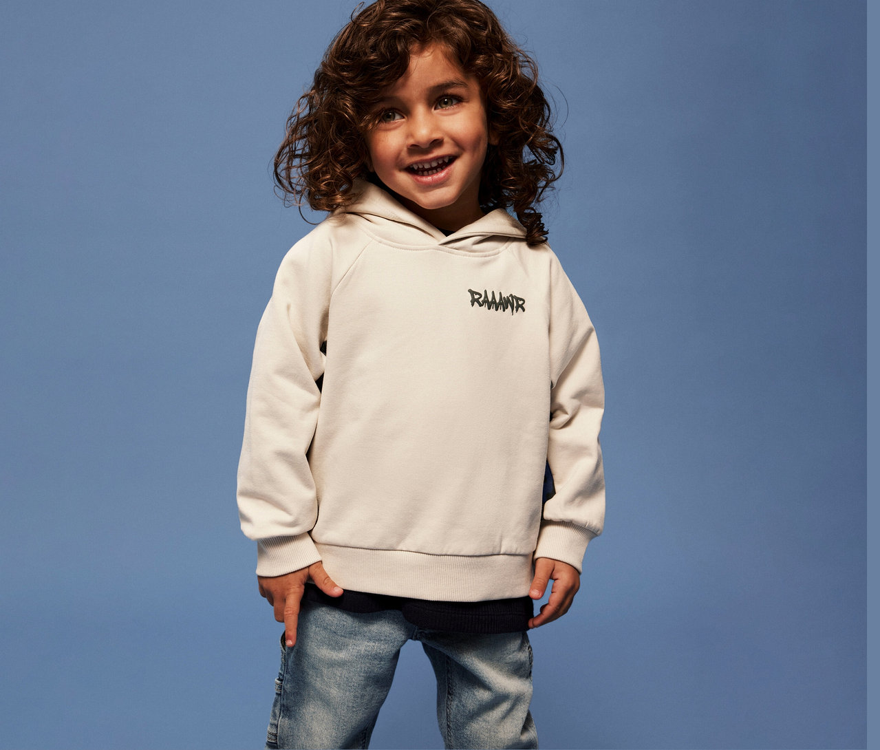 Un garçon souriant aux cheveux bouclés porte un sweat-shirt avec application dino en 3D pour enfant NAME IT et un jean sur fond bleu.