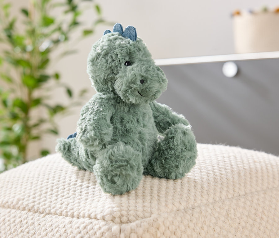 Un dinosaure vert en peluche est assis sur un pouf blanc texturé. Un plante et une armoire sont visibles à l'arrière-plan.