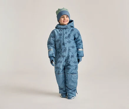 Un garçon souriant porte une combinaison de neige bleue à motifs de dinosaures, des gants assortis et un bonnet sur un fond clair.