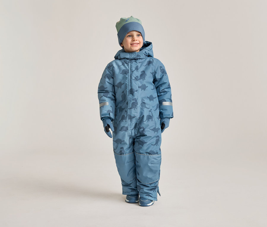 Un garçon souriant porte une combinaison de neige bleue à motifs de dinosaures, des gants assortis et un bonnet sur un fond clair.