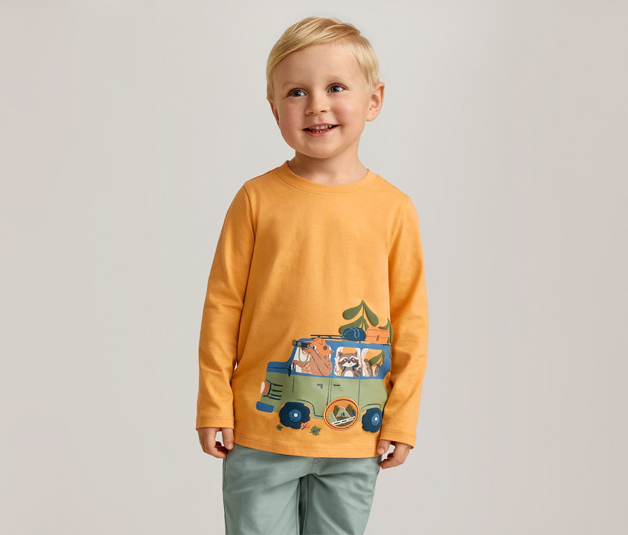 Un garçon souriant aux cheveux blonds porte un t-shirt orange à manches longues avec un imprimé de voiture verte.