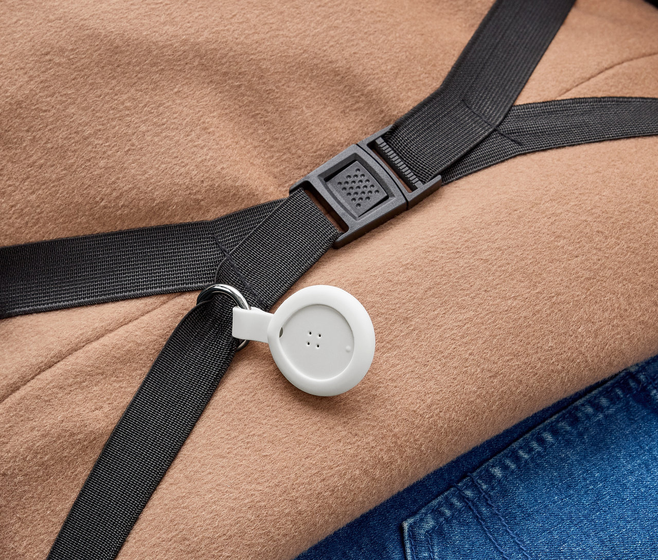 Un appareil blanc et circulaire avec trois points est attaché à une sangle noire avec un anneau métallique. La sangle, dotée d'une boucle en plastique noire, repose sur un tissu marron clair et du denim bleu.