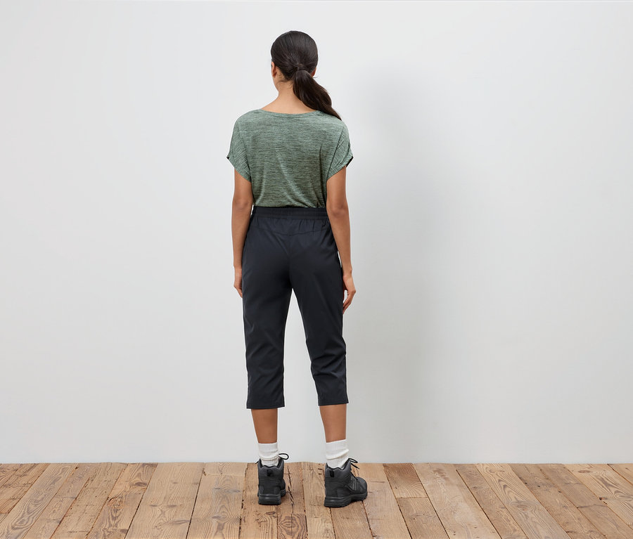 Vue de dos d'une femme portant un t-shirt vert olive, un pantacourt noir et des chaussures de randonnée sur un plancher en bois.