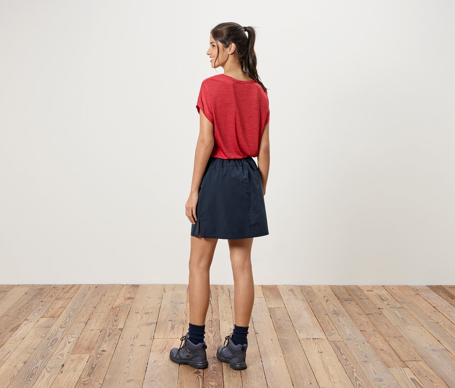 Une femme se tient dos à la caméra, elle porte un t-shirt rouge, une jupe bleue et des chaussures de randonnée grises.