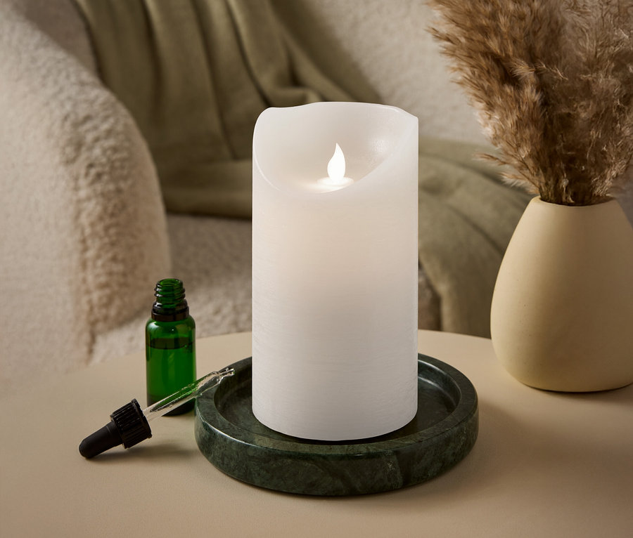 Une bougie pilier blanche avec une flamme LED repose sur un plateau en marbre vert à côté d'une bouteille en verre verte avec une pipette.