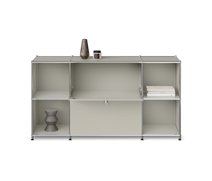 Meuble de rangement modulaire gris clair avec cadre chromé. Un vase foncé et une bougie rose sont posés dessus. À l'intérieur, un objet décoratif gris à gauche, trois livres clairs à droite, et une porte abattante avec serrure au centre.