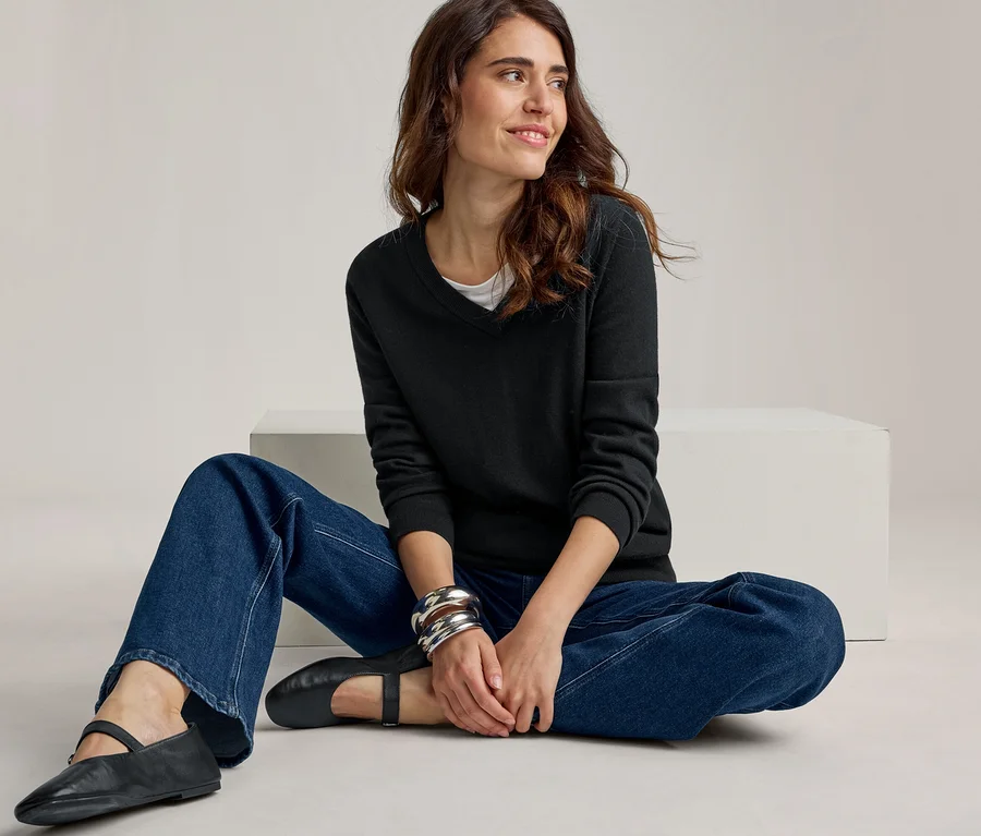 Une femme est assise en tailleur sur le sol, regardant vers la droite avec un léger sourire. Elle porte un pull noir à col en V, un haut blanc, un jean bleu large et des chaussures plates noires. Elle a plusieurs bracelets argentés au poignet gauche.