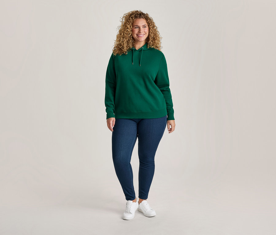 Femme souriante aux cheveux bouclés portant un sweat à capuche vert, un jean bleu et des baskets blanches, posant devant un fond clair.