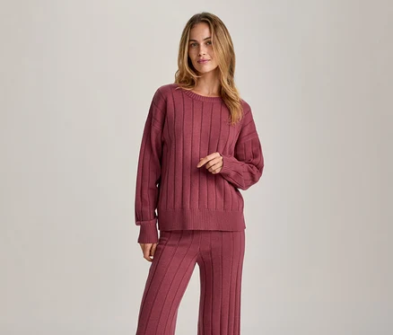 Une femme aux longs cheveux blonds, vêtue d'un pull côtelé à manches longues et d'un pantalon côtelé assorti de couleur rose-rouge, se tient debout et regarde devant elle.
