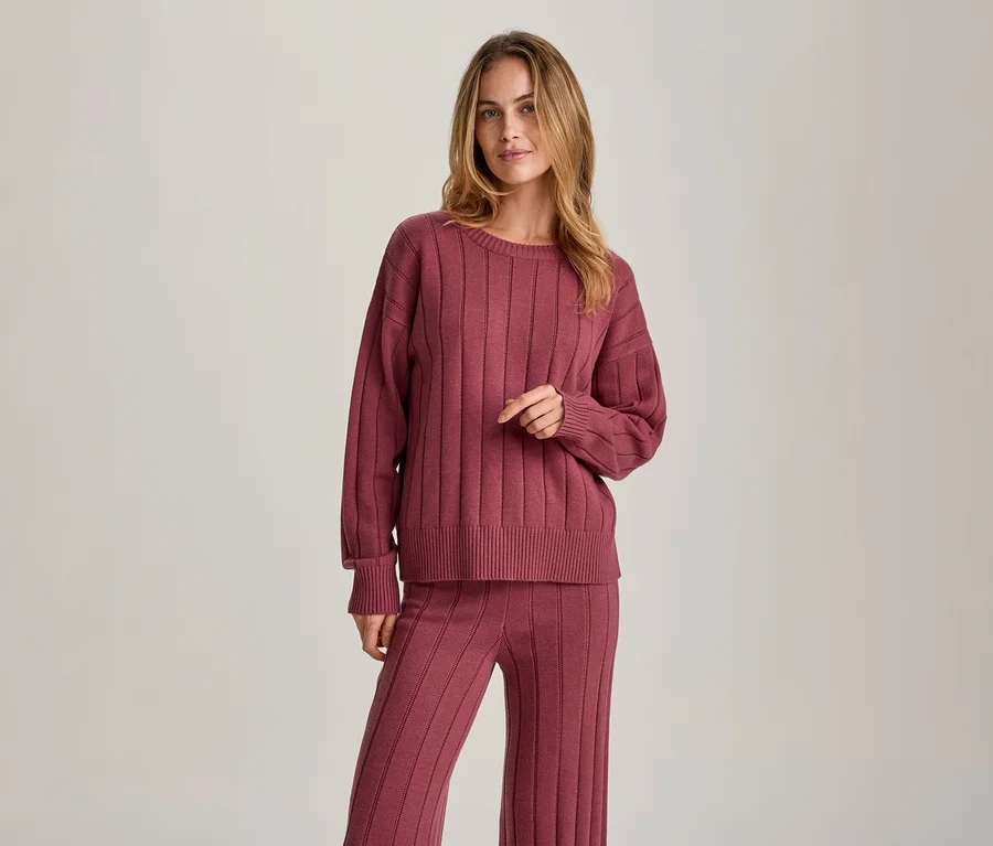 Une femme aux longs cheveux blonds, vêtue d'un pull côtelé à manches longues et d'un pantalon côtelé assorti de couleur rose-rouge, se tient debout et regarde devant elle.