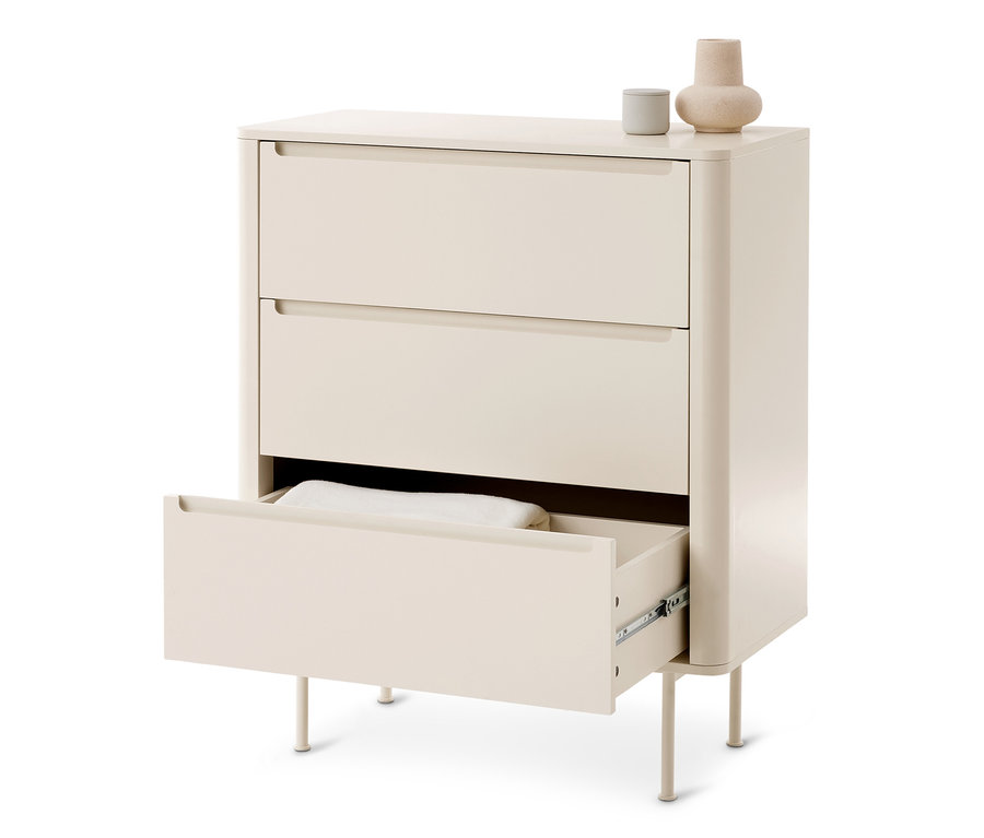 Commode crème clair à trois tiroirs; le tiroir du bas est ouvert et contient un textile blanc. Un petit récipient gris et un vase beige sont posés dessus.