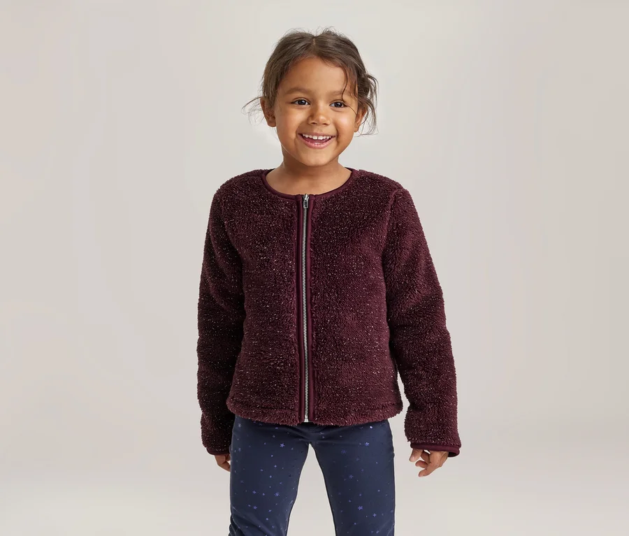 Petite fille souriante portant une veste zippée bordeaux duveteuse et un pantalon bleu foncé à motifs.