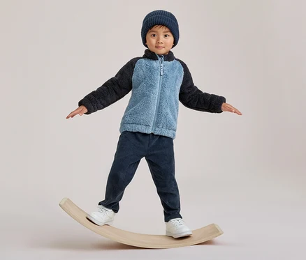 Un enfant en bonnet bleu, veste polaire bicolore et pantalon en velours côtelé bleu foncé, en équilibre sur une planche en bois.