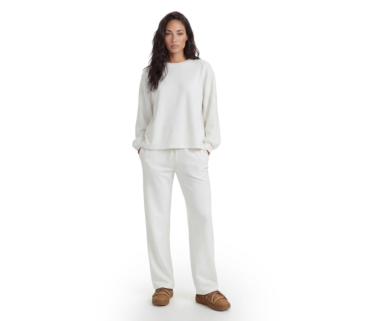 Femme debout, vêtue d'un haut blanc à manches longues, d'un pantalon de survêtement blanc assorti et de chaussons marron.