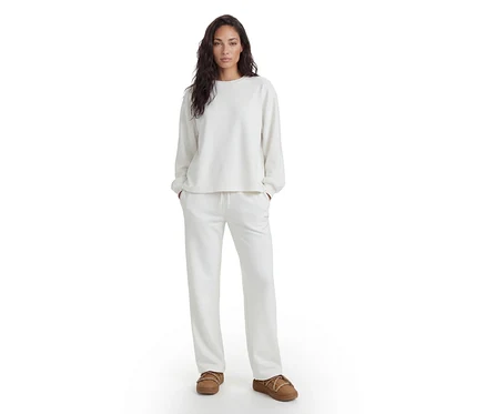 Femme debout, vêtue d'un haut blanc à manches longues, d'un pantalon de survêtement blanc assorti et de chaussons marron.
