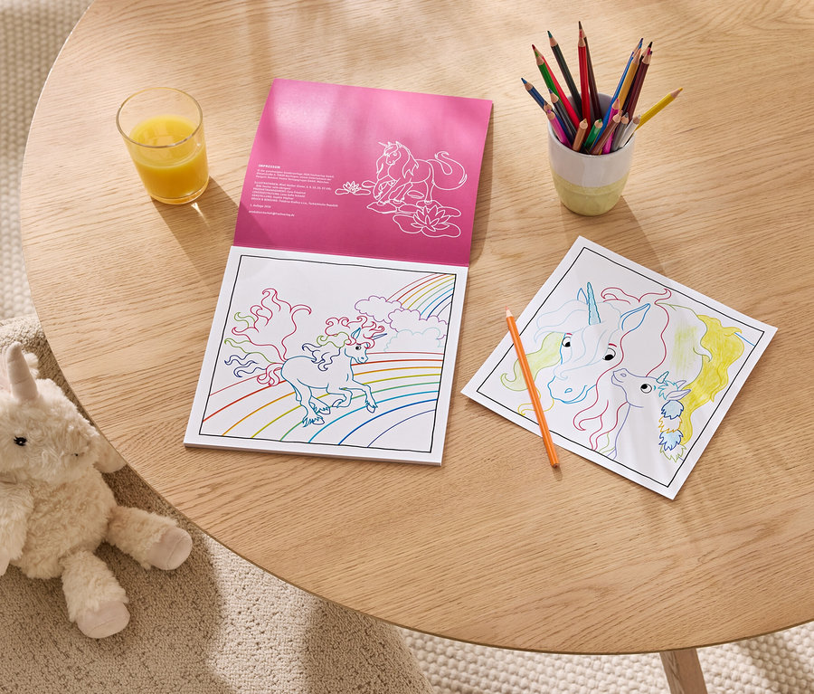 Un livre de coloriage rose licorne est ouvert sur une table en bois, à côté d'une page de coloriage licorne partiellement colorée, d'un pot de crayons de couleur et d'un verre de jus d'orange. Une peluche licorne blanche et moelleuse est assise sur une chaise beige près de la table.