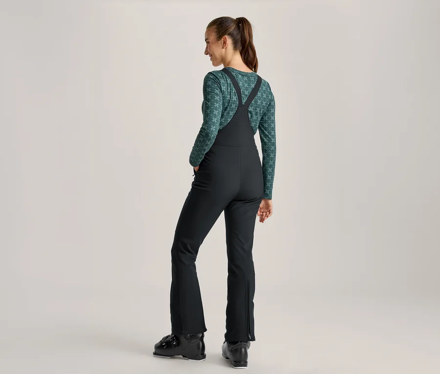 Femme vue de dos en tenue de ski, avec un haut à manches longues vert à motifs, une salopette de ski noire et des chaussures de ski noires. Ses cheveux sont attachés en queue de cheval, elle regarde sur le côté.
