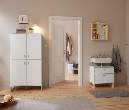 Salle de bain lumineuse avec parquet en bois et murs gris clair. À gauche, une armoire blanche avec une lampe de table à LED sans fil blanche et une plante. À droite, un miroir avec étagères est suspendu au-dessus d'un lavabo avec meuble sous-vasque. Une porte ouverte mène à une pièce adjacente.