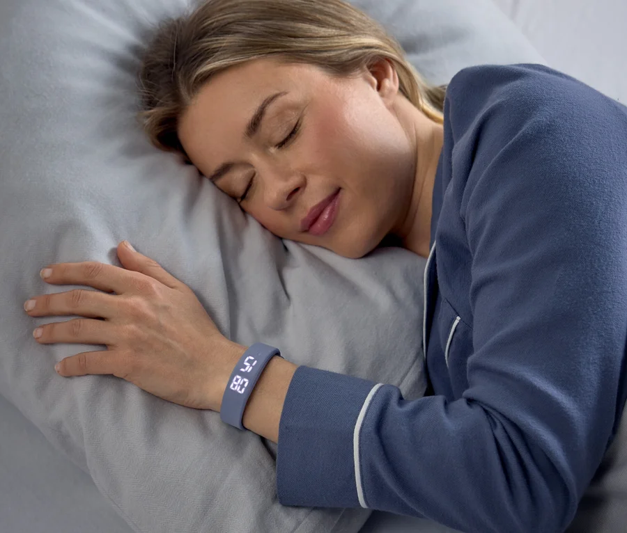 Une femme dort paisiblement sur le côté sur un oreiller gris, vêtue d'un pyjama bleu. Un bracelet connecté gris à son poignet gauche affiche "58" et "80".