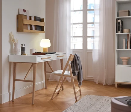 Un bureau blanc avec deux tiroirs, une lampe de table à LED sans fil allumée et un ordinateur portable. À côté se trouve une chaise pliante en bois clair avec une couverture grise. Au-dessus du bureau, une étagère murale en bois est fixée.