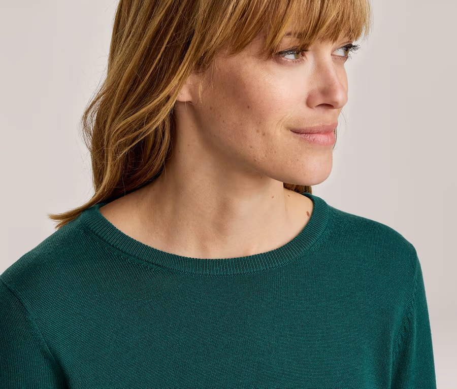 Gros plan d'une femme aux cheveux châtains clairs avec une frange, portant un pull vert foncé à col rond et regardant vers la droite.