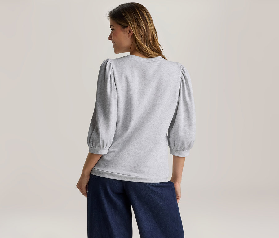 Femme vue de dos, portant un haut gris clair à manches bouffantes trois-quarts et un pantalon large bleu foncé, regardant vers sa droite.