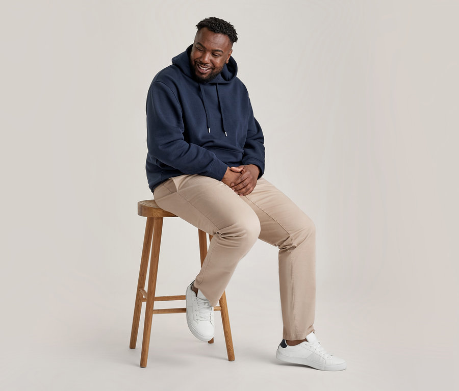 Un homme souriant avec une barbe est assis sur un tabouret en bois, portant un sweat à capuche bleu marine, un pantalon chino beige et des baskets blanches.