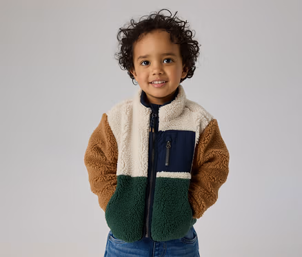 Enfant souriant aux cheveux bouclés portant une veste polaire color-block crème, marron, vert foncé, avec une poche poitrine bleu marine.