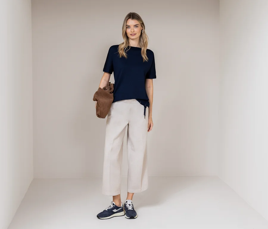 Une femme se tient dans une pièce, portant un t-shirt bleu marine, un pantalon beige et des baskets bleues. Elle tient un sac marron.