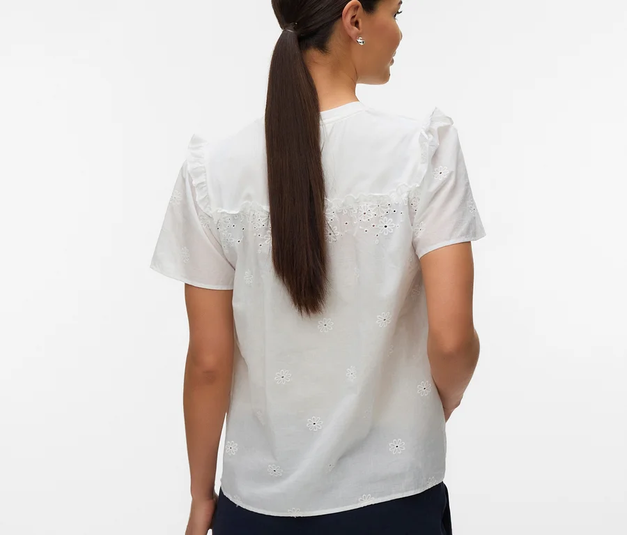 Une femme vue de dos, légèrement tournée, porte un haut MAMALICIOUS « MLDORA » blanc à manches courtes avec des détails brodés, associé à un pantalon bleu foncé. Ses cheveux foncés sont attachés en longue queue de cheval.