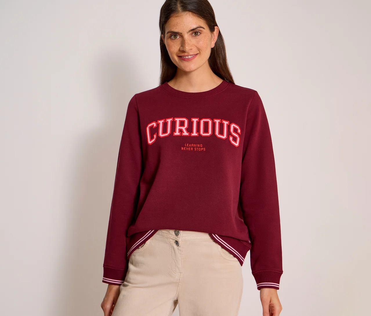 Une femme souriante aux cheveux foncés porte un sweatshirt CECIL bordeaux avec l'inscription « CURIOUS » en lettres rouges et blanches et « LEARNING NEVER STOPS » en dessous, associé à un pantalon beige clair.