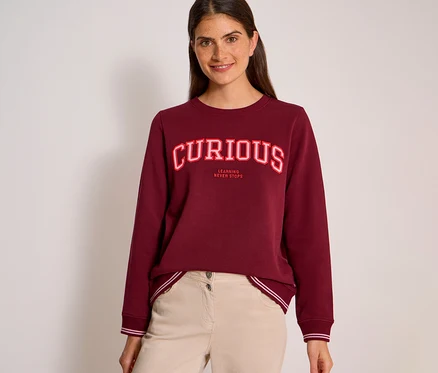 Une femme souriante aux cheveux foncés porte un sweatshirt CECIL bordeaux avec l'inscription « CURIOUS » en lettres rouges et blanches et « LEARNING NEVER STOPS » en dessous, associé à un pantalon beige clair.