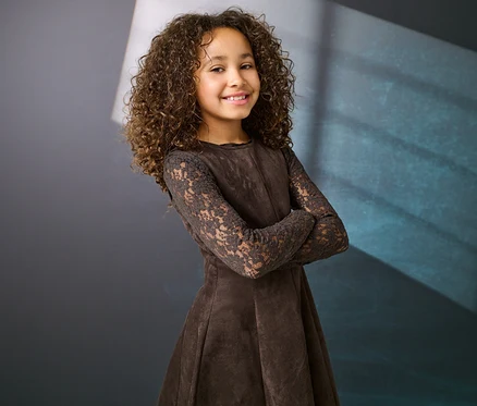 Jeune fille souriante aux cheveux bruns bouclés, vêtue d'une robe marron à manches en dentelle, les bras croisés devant un fond sombre.