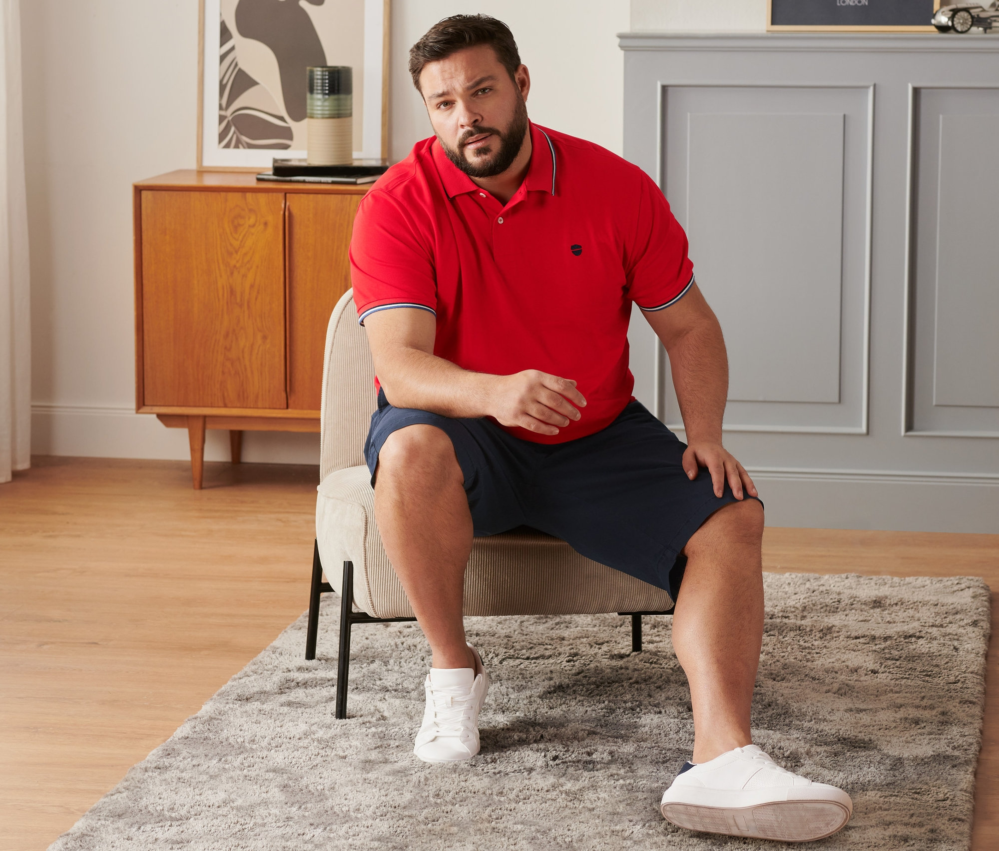 Un homme est assis sur une chaise et porte un short chino bleu marine, un polo en piqué rouge et des tennis.
