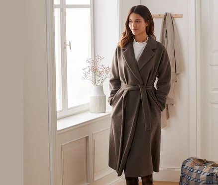 Une femme porte un manteau en laine mélangée et se tient près d'une fenêtre.